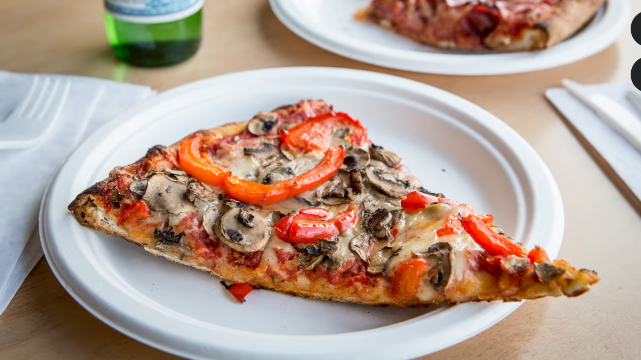 A slice of pizza topped with red peppers and mushrooms, served on a disposable plate with a second pizza slice in the background.
